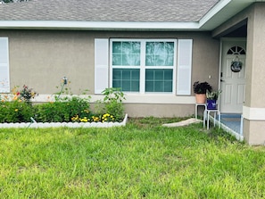 Une maison avec un jardin devant, une porte ornée d’une guirlande et un pot de fleurs posé sur un support.