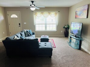 A living room with a sofa, ottoman, TV, ceiling fan, window with curtains, and a painting on the wall.