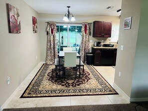 Un coin repas avec une table et des chaises, un tapis à motifs, une cuisine avec des placards et une fenêtre avec des rideaux.
