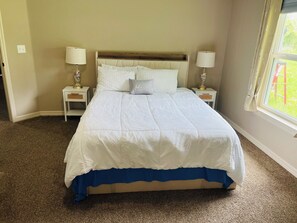 Un lit bien fait, avec une couvre-lit bleu, deux tables de chevet avec des lampes, une fenêtre avec des rideaux et un sol recouvert d’une moquette.