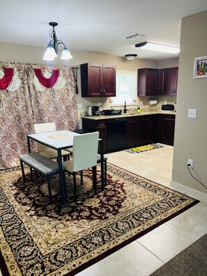 Un comedor con mesa y sillas, una cocina con armarios oscuros, un tapete estampado y una lámpara en el techo.