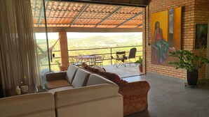 A spacious living area with a large sofa, a dining table with chairs, and a balcony with a view of the hills.