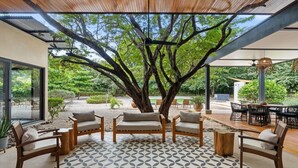An outdoor seating area with wooden furniture and a patterned floor, surrounded by trees and a view of a garden. - Villa in Exclusive Community 3-Min Drive to Beach w/ Private Pool, Sauna & Cold Plunge (Hacienda Pinilla)