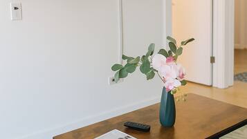 Un salon avec un téléviseur à écran plat fixé au mur, une table basse avec une télécommande, un vase rempli de fleurs et un magazine.