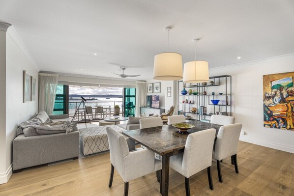 A modern living room with a dining area, large windows, and a view of the sea.