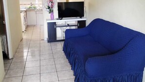 A living room with a blue sofa, a flat-screen TV on a stand, and a kitchen in the background.