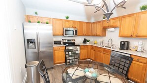 A compact kitchen with wooden cabinets, stainless steel appliances, a glass dining table, and a modern chandelier.