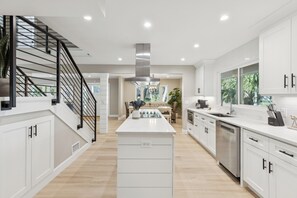 A modern kitchen with white cabinetry, a central island, stainless steel appliances, and a staircase with black railings. - 4-Bedroom beach house, steps away from Coligny Plaza (Hilton Head Island)