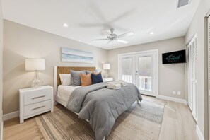 Une chambre à coucher avec un lit, des tables de chevet, un ventilateur de plafond, une télévision et une vue sur l’extérieur.
