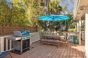 Une terrasse extérieure équipée d’un barbecue, d’une table en bois, de bancs et d’un parasol bleu.