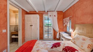 Ein Schlafzimmer mit einem Bett, einem hölzernen Kopfteil, einem Fenster mit Vorhängen, einer Tür und einem kleinen Tisch mit Lampe.