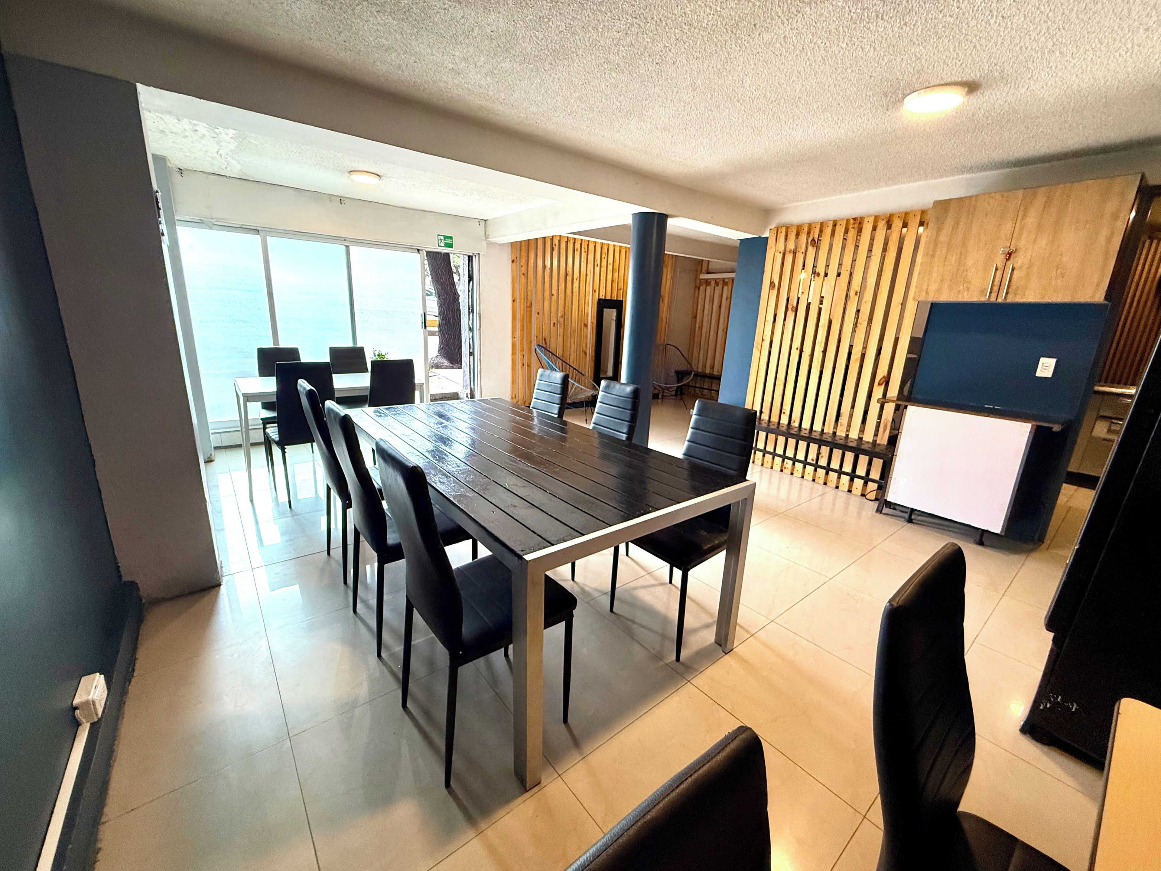 A modern dining area with a long table and chairs, a wooden accent wall, and a small kitchenette.