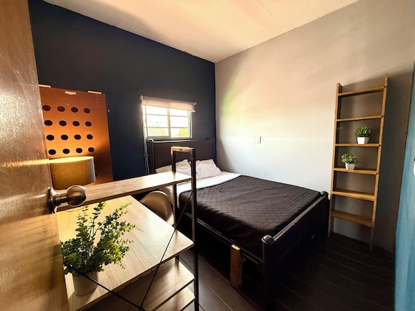 A compact room with a bed, a desk, a chair, a window, and a shelving unit. - RHM Spaces - Santa Maria (Mexico City)