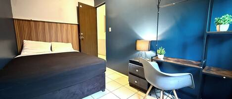 A bedroom with a bed, a desk with a chair, a lamp, and a shelf with a plant.