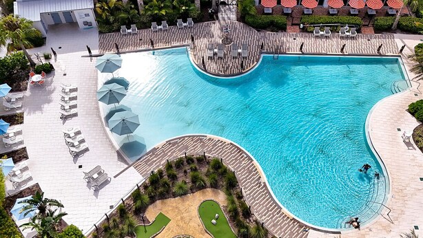 A large, curved swimming pool with lounge chairs and umbrellas around it, surrounded by greenery and a paved walkway.