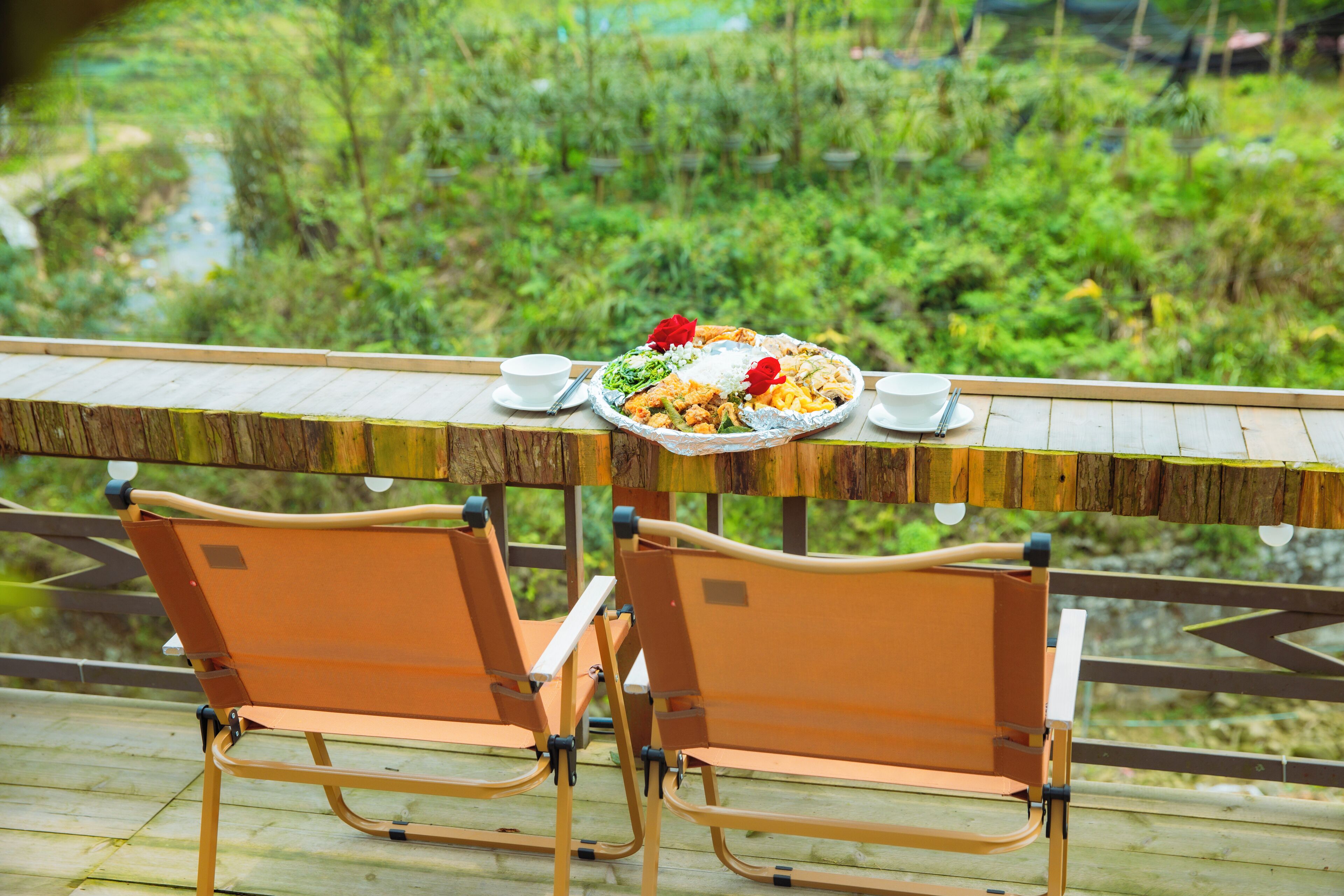 A dining table with a meal and two chairs on a wooden deck with a view of greenery.