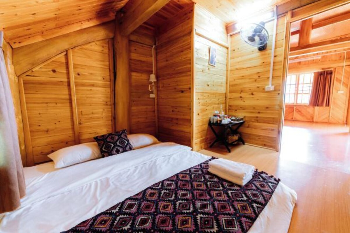 Un dormitorio de cabaña de madera con cama, almohadas, una mesita con objetos y una ventana.
