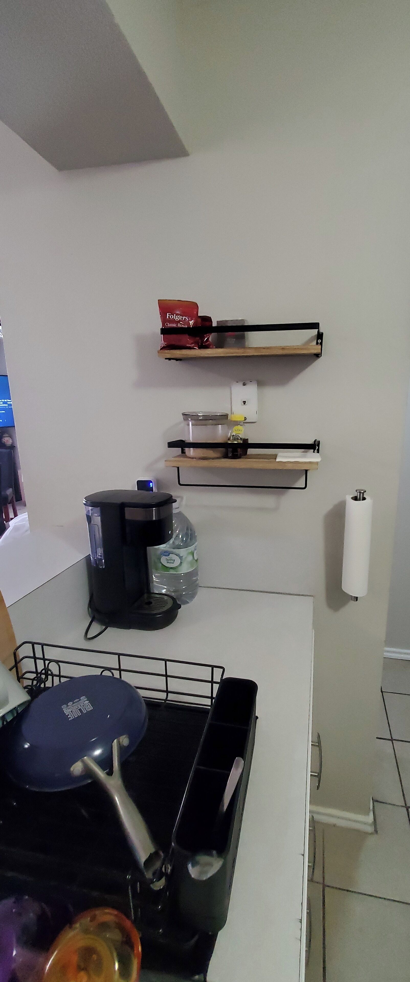 A kitchen with a coffee maker, a kettle, a frying pan, and a shelf with a red box.