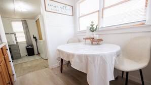 Un comedor con una mesa redonda cubierta con un mantel blanco, una silla blanca y una mesita de madera con un florero y una vela.