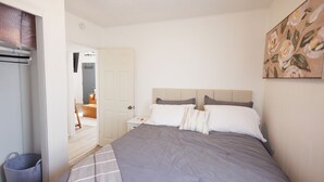 Un dormitorio con una cama grande, una mesita de noche, un cuadro colgado en la pared y una puerta que da a otra habitación.