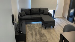 Un salon moderne avec un canapé d’angle, une petite table d’appoint, un tapis et un coin cuisine équipé d’un réfrigérateur.