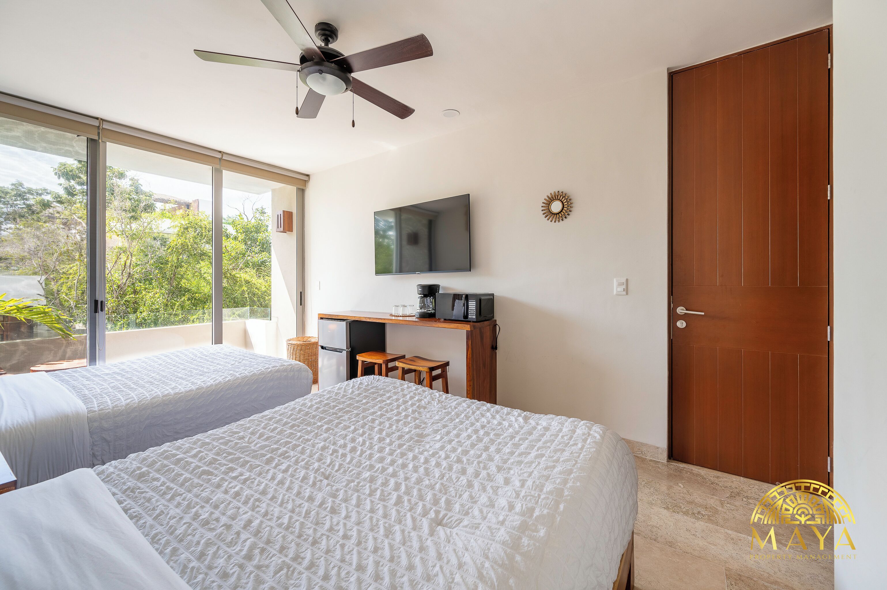 A hotel room with two beds, a desk with a TV, a ceiling fan, and a large sliding door.