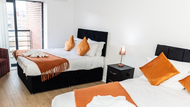 A modern hotel room with two beds, a black headboard, and a nightstand with a lamp. - Apartment Sleeps 6 with Balcony and Parking (Birmingham)