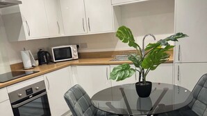 A modern kitchen with white cabinets, a microwave, and a coffee maker. There is a round glass table with two chairs and a potted plant.