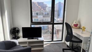 A modern hotel room with a large window, a flat-screen TV, a desk, a chair, and a bed.