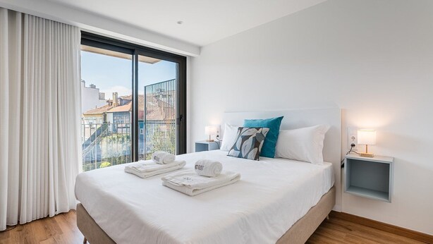Een moderne hotelkamer met een groot bed, twee nachtkastjes met lampen, uitzicht op gebouwen door een balkondeur, en een net opgemaakt bed met handdoeken.