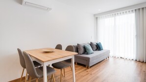 Een moderne woonkamer met een eettafel, een grijze bank met kussens en een groot raam met dunne gordijnen.