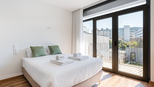 A modern hotel room with a large bed, green pillows, white linens, and a balcony with a city view. - GuestReady Essential - The Gods: Baldur (Porto)