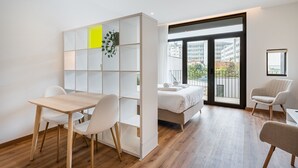 A modern hotel room with a wooden floor, a white bed, a desk with chairs, a balcony with a view of buildings, and a large window. - GuestReady Essential - The Gods: Baldur (Porto)
