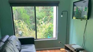 Una habitación con sofá de cuero negro, suelo de madera, ventana grande con vista a follaje y televisor montado en la pared.
