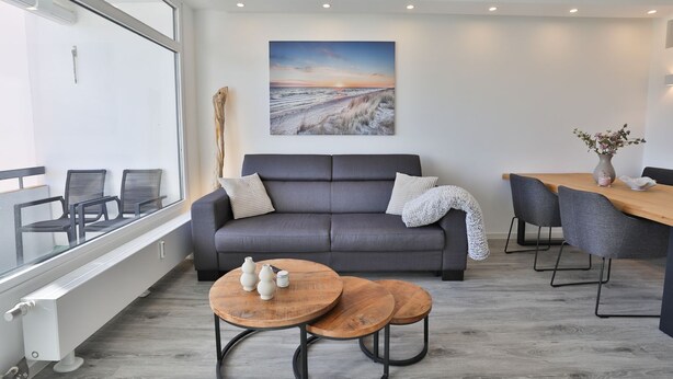 A modern living room with a grey sofa, a wooden coffee table, and a dining area with a wooden table and chairs. - Strandkoje 104 - Holiday apartment near the beach in Scharbeutz - Modern, quiet & sunny (Scharbeutz)
