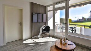 A modern room with a large window, a flat-screen TV, a chair, a round wooden table, and a potted plant. - Strandkoje 104 - Holiday apartment near the beach in Scharbeutz - Modern, quiet & sunny (Scharbeutz)