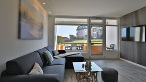 A modern living room with a grey sofa, a glass coffee table, and a flat-screen TV mounted on a wall with vertical lines. - Strandkoje 105 - Modern vacation apartment in Scharbeutz - Close to the beach, quiet & sunny (Scharbeutz)