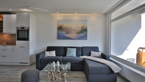 A modern living room with a grey sectional sofa, a glass coffee table, and a painting of a beach scene. - Strandkoje 105 - Modern vacation apartment in Scharbeutz - Close to the beach, quiet & sunny (Scharbeutz)