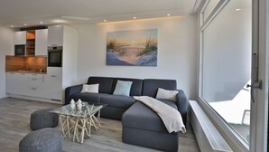A modern living room with a grey sofa, a glass coffee table, and a painting of a beach scene. - Strandkoje 105 - Modern vacation apartment in Scharbeutz - Close to the beach, quiet & sunny (Scharbeutz)