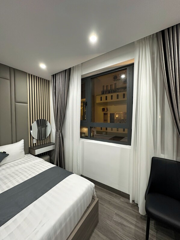 A modern hotel room with a large bed, a bedside table with a mirror, a window with curtains, and a view of buildings outside.
