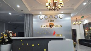 Un comptoir d'accueil d'hôtel au design moderne, agrémenté d'un lustre, d'horloges indiquant l'heure dans différentes villes et d'une enseigne indiquant le nom de l'hôtel : « SEVEN STARS ».