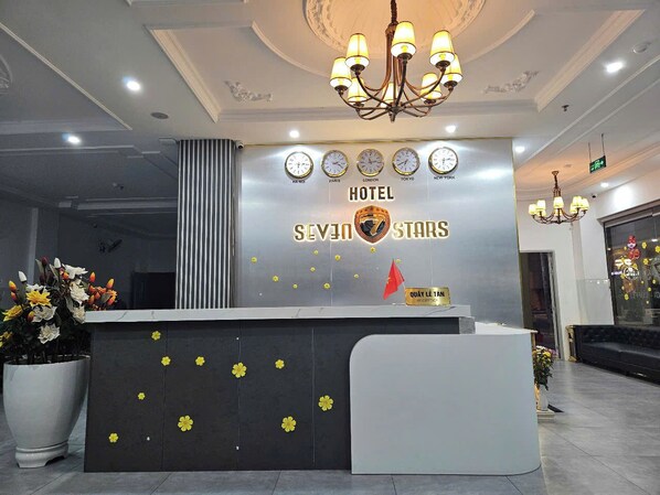 Un mostrador de recepción de hotel con diseño moderno, que incluye un candelabro, relojes mostrando la hora en varias ciudades y un cartel con el nombre del hotel 'SEVEN STARS'.