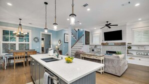 A modern kitchen with a central island, a dining area with wooden chairs, a grey sofa, and a staircase leading to another level. - 65 Water Oak Way-Ocean Oaks (Jekyll Island)