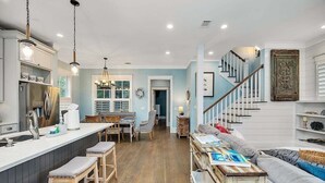 A spacious kitchen with a central island, a dining area with chairs, a staircase leading to a loft, and a living area with a sofa and cushions. - 65 Water Oak Way-Ocean Oaks (Jekyll Island)