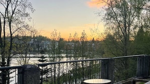Ulkona on oleskelualue, jossa on pöytä ja penkkejä. Alueelta avautuu näkymä joelle, ja sitä ympäröivät puut sekä kaide.