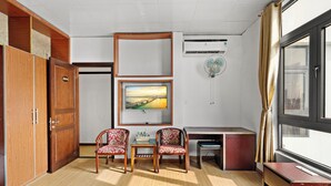 Ein Zimmer mit einem Holzschrank, zwei gemusterten Stühlen, einem kleinen Tisch mit Flaschen, einem Schreibtisch und einem großen Fenster mit Vorhängen.