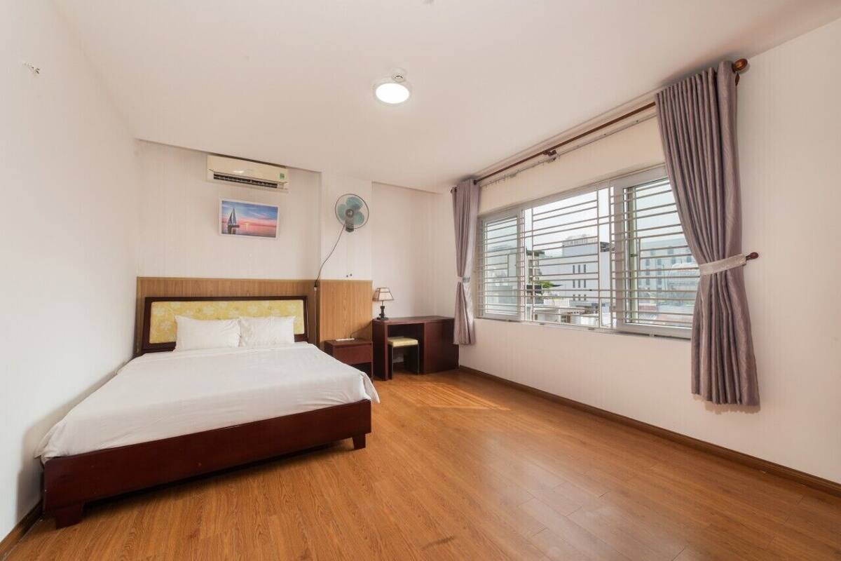 Een slaapkamer met een houten bed, een bureau, een stoel, een ventilator en een raam met gordijnen.
