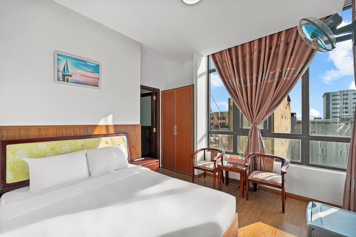 Een hotelkamer met een groot bed, een houten garderobe, een bureau met stoel, en een uitzicht op de stad door grote ramen.