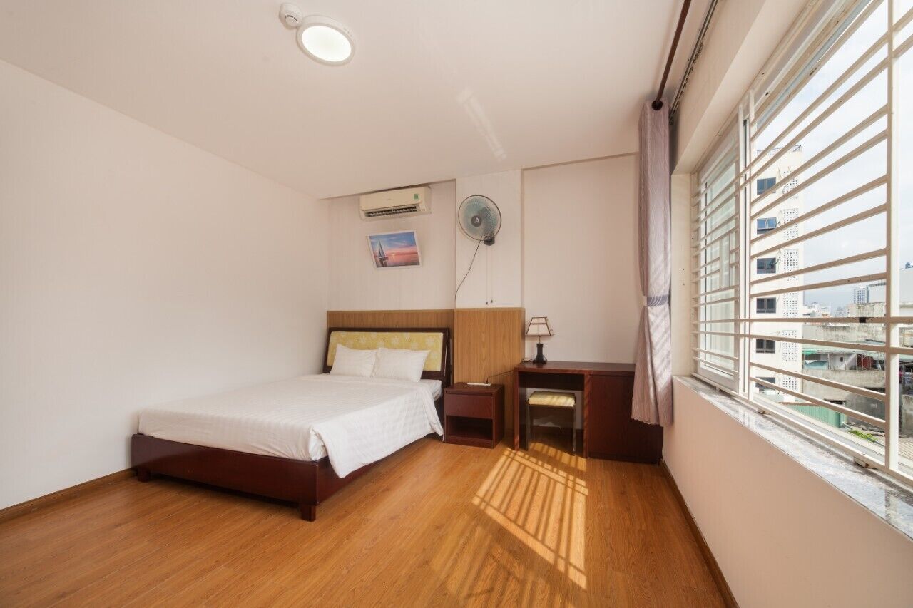 A bedroom with a bed, bedside table, lamp, and a view of buildings through the window.