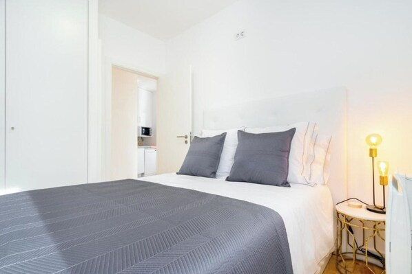 A neatly made bed with a gray bedspread and pillows, a bedside table with a lamp, and a small kitchenette in the background.